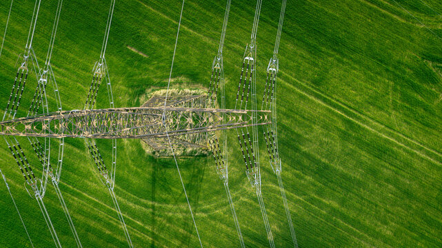 Aerial top-down view of a high-voltage power line pylon in a vibrant green agricultural field. Industrial infrastructure meets nature in a symmetrical composition.