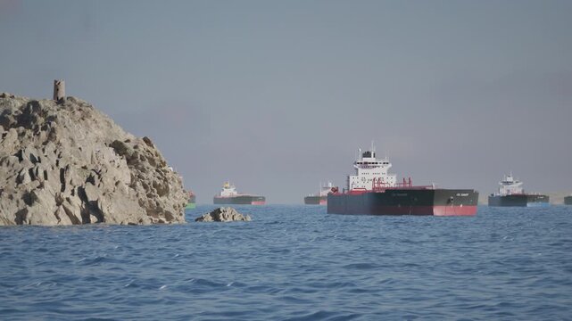 Camera pans along stony Headland to reveal Crude Oil Tanker Ships anchored in Bay. Aerial View of Oil Tankers anchored in the Ocean. Strait of Hormuz Blockade Concept. 3d Animation Render