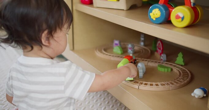 A young toddler develops fine motor skills through imaginative play, pushing a wooden train along a track on a shelf in a modern nursery environment.