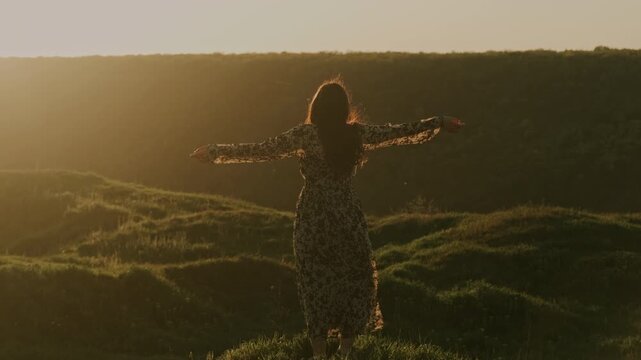 Woman Raises Arms At Sunset, Silhouetted On Grassy Ridge With Warm Backlight, Long Dress Fluttering, Golden