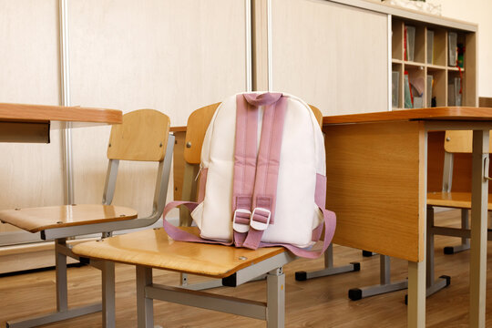 Pink schoolbag on wooden chair in empty classroom. Backpack for girl on desk background in school class. Back to school concept, elementary education, student supplies, learning furniture, nobody