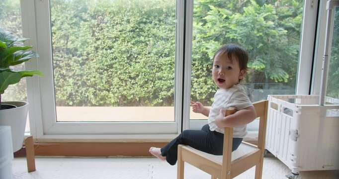 A curious Asian toddler sits on a wooden chair in a sunlit room, observing her environment in a moment of quiet learning and early childhood development.