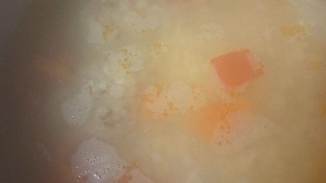 Cooking process of vegetable soup with carrots and potatoes simmering in a pot, bubbles forming on the surface, steam rising in a warm kitchen setting