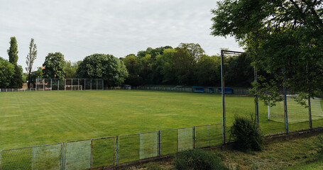 Empty football field with green grass and goal posts surrounded by dense trees under cloudy daylight. Outdoor sport, recreation, training ground, and community leisure concept. © Ekaterina