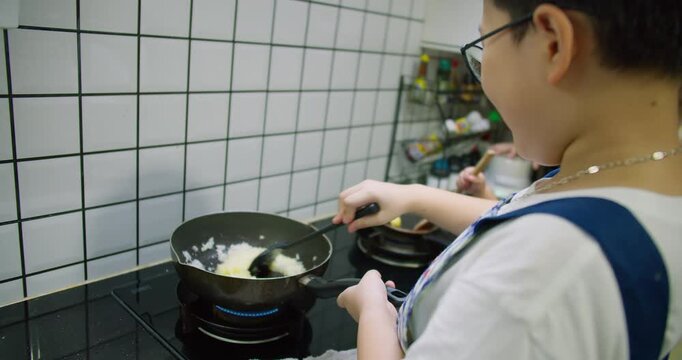 A young boy learning to cook stirs ingredients in a frying pan on a modern gas stove, developing practical life skills through a hands-on home cooking lesson.