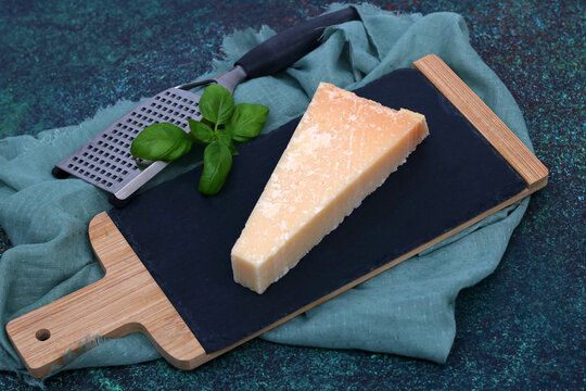 A piece of Parmesan hard cheese with a cheese grater and basil on a rustic board.