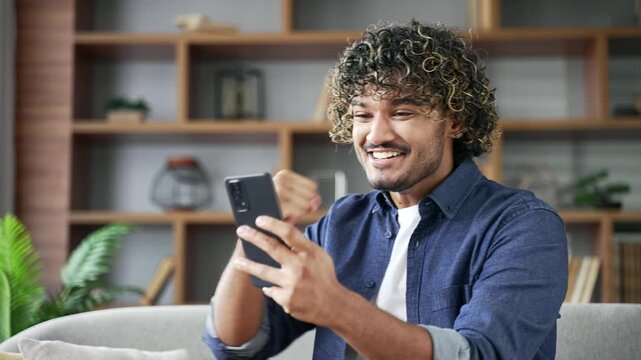 Happy excited man received great news on the mobile phone while sitting on sofa in living room at home. Smiling satisfied handsome male reads a positive good message, celebrates success. Close up