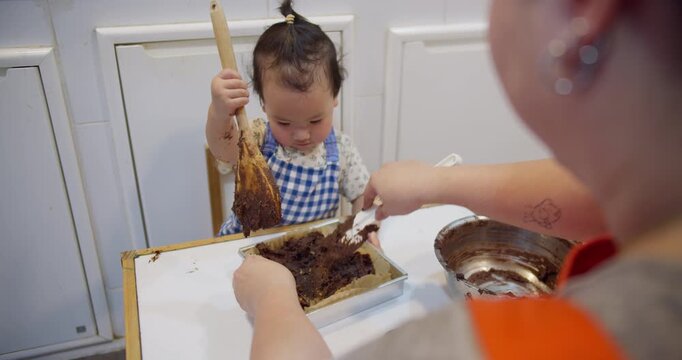 A focused toddler wearing an apron learns to bake chocolate batter with family, engaging in a fun, educational sensory play activity together in the kitchen.