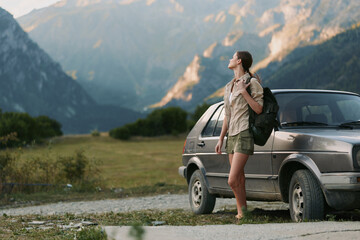 Obraz premium Young woman traveler with backpack stands near car on mountain road looks at scenic landscape enjoys nature and outdoor adventure lifestyle in summer day.