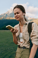 Naklejka premium Smiling woman outdoors checking smartphone during a hike. Casual outfit and backpack against a mountainous landscape. The scene conveys lifestyle, exploration, and wellness.