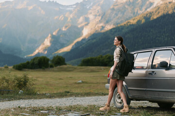 Naklejka premium Energetic woman outdoors beside a car, exploring mountain scenery, sunlit moment, casual travel gear, serene atmosphere, nature connection, fresh air and freedom in nature under blue sky