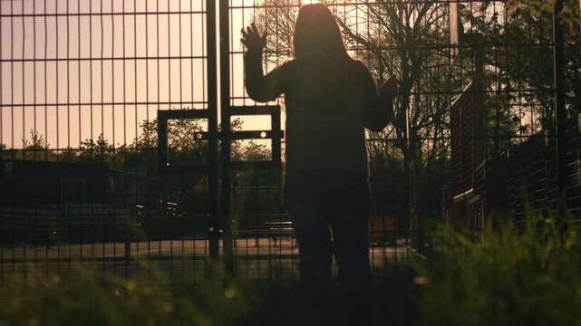 Mother waiting at school gates with playground at dusk 