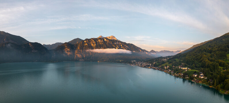Aerial panorama of Attersee lake in Austria at summer sunrise, with alpine mountains, mist, forested hills and a peaceful lakeside village.
