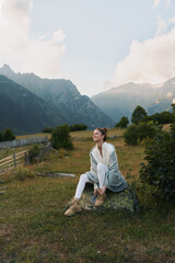 Naklejka premium Young woman enjoys outdoor lifestyle in a mountain meadow, sits on a rock, wearing light cardigan and boots, tranquil landscape behind, calm mood and natural surroundings