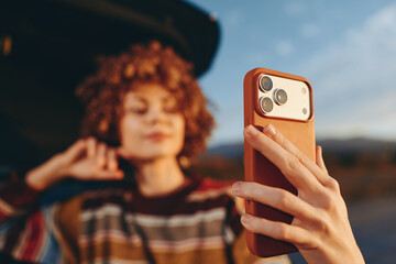 Woman lifestyle portrait outdoors, holding smartphone for a selfie, hand near face, curly hair...