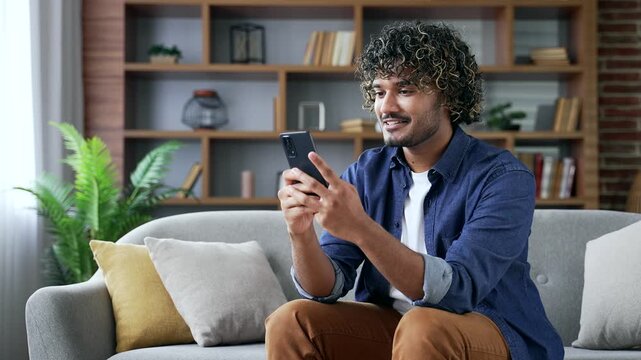 Portrait of happy young handsome man holding mobile phone sitting on sofa in living room at home. Confident smiling male using smartphone at leisure. Positive friendly male poses looking at camera