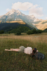 Naklejka premium Calm woman lying in a sunlit meadow with distant mountains, relaxed pose, gentle breeze, natural colors, lush grass, outdoor lifestyle moment soft light, open landscape.