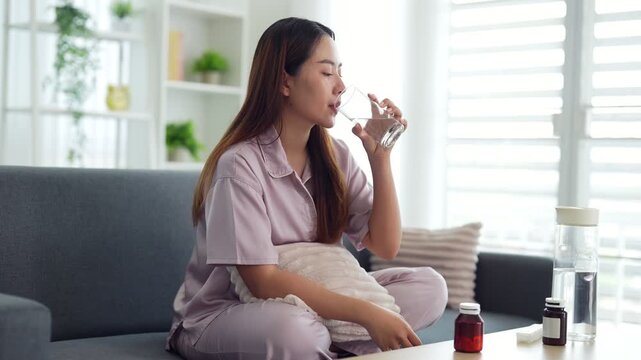 young Asian women take medicine pill drink water at home sit on sofa living room morning healthcare wellness treatment sick patient medical vitamin supplement