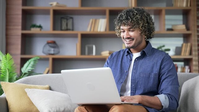 Satisfied happy young man received great news looking at laptop screen sitting on sofa in living room at home. Smiling glad handsome male freelancer reads a pleasant message on computer, rejoices