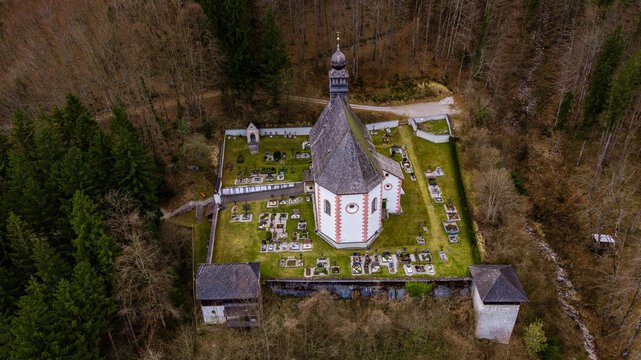 Aerial view of the historic Bergkirche Klaus church and its walled cemetery surrounded by dense forest and a small stream in Klaus an der Pyhrnbahn, Upper Austria, Austria.
