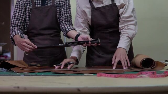 Two hands measuring leather on table with tape and ruler, apronclad collaborators marking belt strap layout, mentor and apprentice