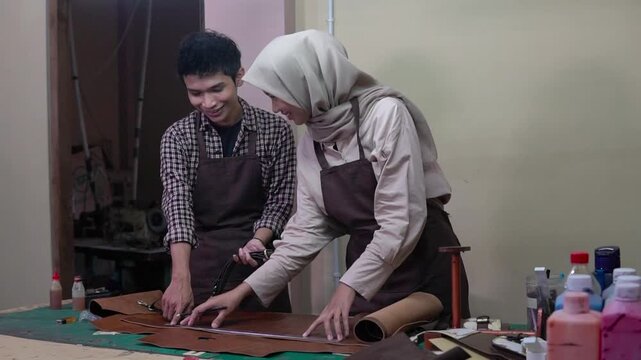 Two asian artisans cutting leather in studio, female wearing hijab guiding male apprentice, measuring pattern on brown hide,