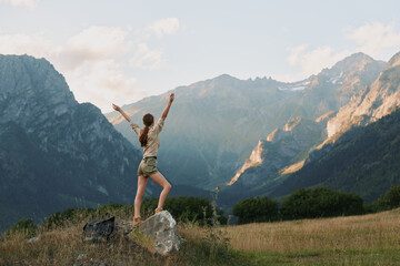 Fototapeta premium Cheerful woman outdoors, standing on a rock with arms raised, mountain valley backdrop, sunny day, carefree mood, nature inspired lifestyle