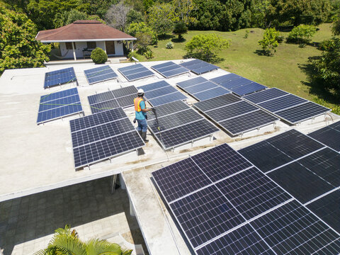 Aerial view of a worker in a safety vest cleaning solar panels on a rooftop surrounded by lush tropical greenery in Guanaja, Bay Islands Department, Honduras.
