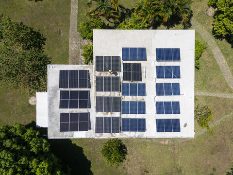 Aerial view of solar panels installed on a white flat roof with a technician working, surrounded by lush green lawns and tropical trees in Guanaja, Bay Islands Department, Honduras.