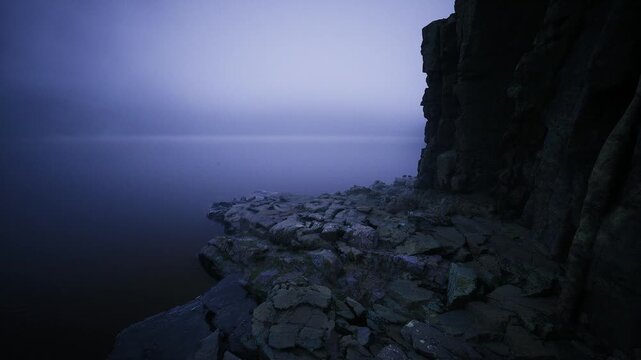 Uneven rocky forecliff leading to calm sea, jagged ledges and slick stone surfaces descend to still water, muted blue tones and tactile textures emphasize