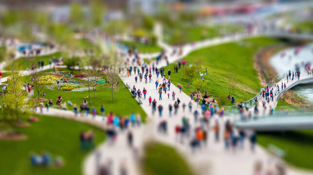 Miniature tilt-shift effect of a busy city park in summer or spring day. People walking on sunny pathways surrounded by green grass and trees. Urban life, summer leisure, tiny toy world perspective.