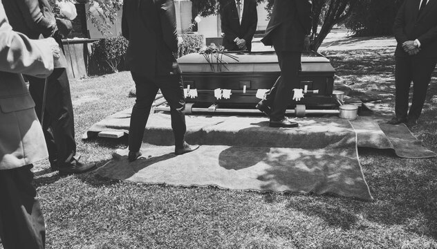 A black and white image of a group of men in suits walking by a casket with flowers and pallbearer gloves at a gravesite