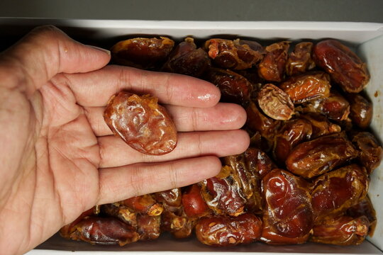 Close-up of a hand holding a single pitted date, split open, showing the internal texture, over a mass of whole dried dates in a simple cardboard box.