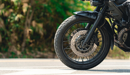 Close-up of a motorcycle front wheel with disc brake on a road, featuring detailed tire treads and spokes against a blurred natural background. © PIPAT