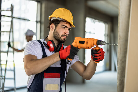 Skilled builder using electric drill on wall with colleagues working in background.
