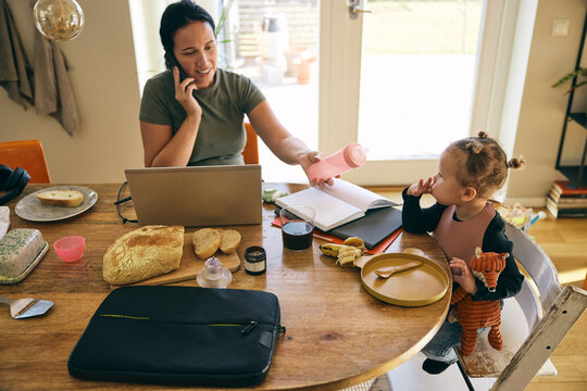 High angle view of freelancer female talking on smart phone and giving feeder to daughter while working from home