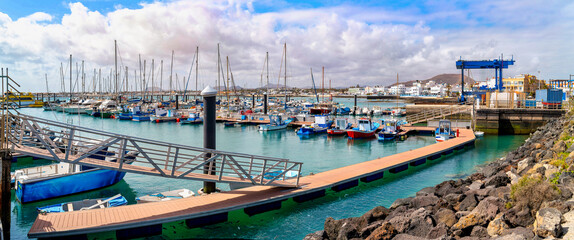 Harbor of Corralejo on Fuerteventura with pier, fishing boats and yachts, turquoise water and coastal town in background under cloudy sky of Canary Islands. © EKH-Pictures
