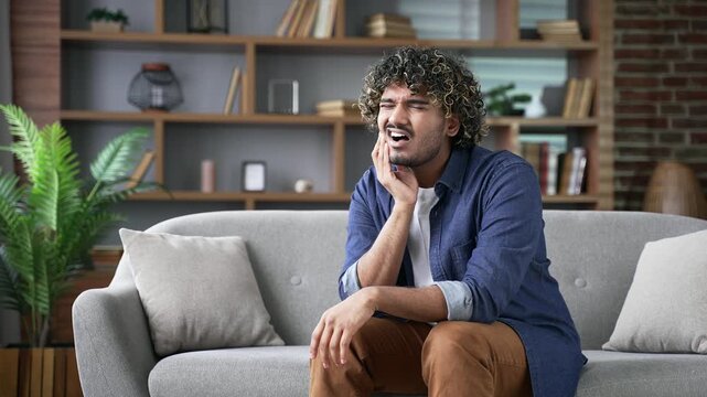 Upset young adult man suffering from toothache while sitting on the sofa in the living room at home. Disappointed male holds his hand near his cheek, massaging a sore spot. He needs help of a dentist