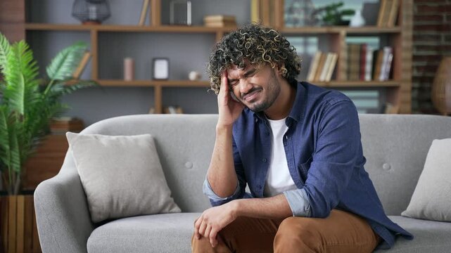 Young adult man suffering from headache while sitting on sofa in living room at home. Upset male massages his temples with his hands. He is tired and feels pain has high blood pressure or migraines