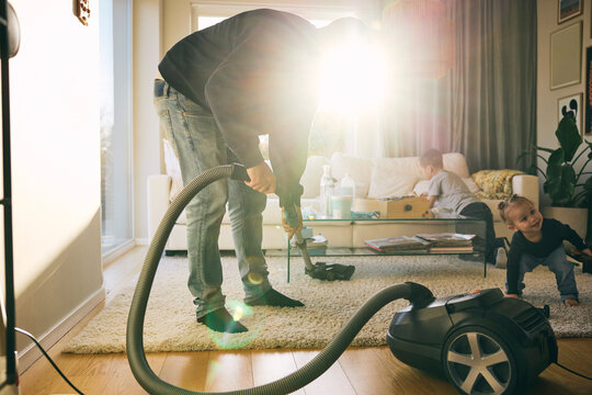 Bright sunlight on man cleaning carpet with vacuum cleaner while kids playing in living room at home