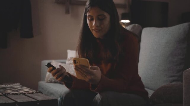 Young woman at home at night switching TV channels with a remote while focused on her smartphone. Multitasking and digital distraction concept, video clip
