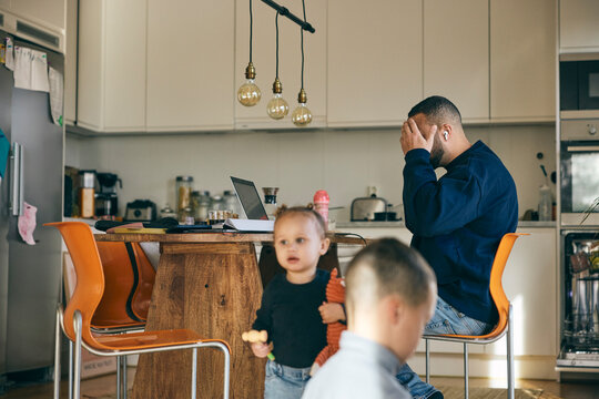 Children playing at home while stressed father working on laptop