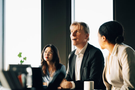Mature male investor in blazer sharing business ideas while sitting with team in office meeting room