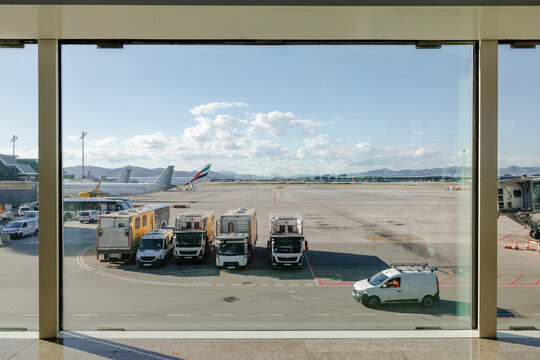 Airport terminal window view in Barcelona at sunset, ground service vehicles and airplanes on runway with mountains in background under sunny spring sky, travel and aviation scene in Europe.