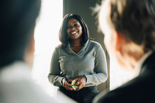 Smiling businesswoman holding adhesive notes and pen while giving presentation to team at office