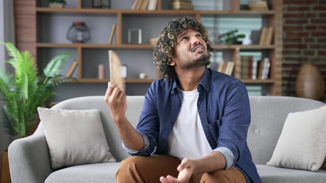 Young adult man is hot while sitting on the sofa in the living room at home. Upset disappointed male waves a fan, feels discomfort and sweating, because the air conditioner in the apartment is broken
