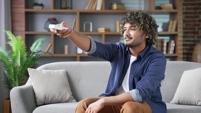 Happy young adult man turns on air conditioner by remote control sitting on sofa in living room at home. Handsome smiling male presses the button of the device and turns off the heating in the house