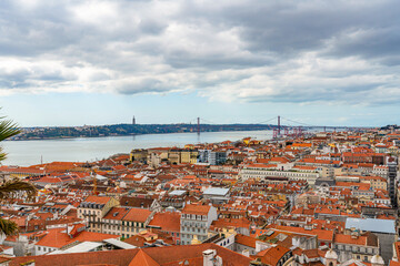 Obraz premium Panoramic view of Lisbon cityscape with red tiled rooftops, Tagus river and 25 de Abril bridge. Iconic urban landscape of Portugal with historic architecture and waterfront skyline under cloudy sky.