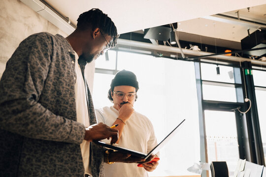 Male computer programmers discussing ideas while working on laptop at modern startup office