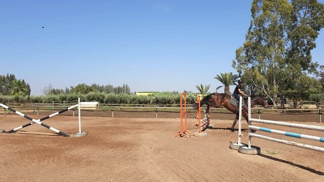 Close-up of a bay horse and rider jumping over a fence in an outdoor equestrian arena. The rider, dressed in black athletic gear and a helmet, 